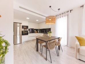 Una cocina moderna con electrodomésticos de acero inoxidable, un comedor con una mesa oscura y sillas de mimbre, y un sofá de color claro con un cojín amarillo.