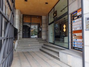 Entrada de un edificio con una escalera que lleva a una puerta de cristal, una valla metálica a la izquierda y un cartel con varios nombres de empresas a la derecha.