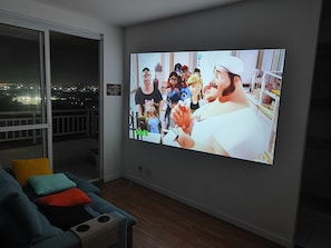 A living room with a large flat-screen TV mounted on the wall, a sofa with cushions, and a wooden floor. - Tricolor Zone! Cozy apartment next to the Grêmio Arena, near the airport. (Porto Alegre)