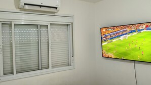 A room with a window featuring horizontal blinds and a mounted flat-screen TV displaying a soccer match. - Tricolor Area! Cozy apartment next to Grêmio Arena, near the airport. (Porto Alegre)