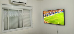 A room with a window featuring horizontal blinds and a mounted flat-screen TV displaying a soccer match. - Tricolor Area! Cozy apartment next to Grêmio Arena, near the airport. (Porto Alegre)