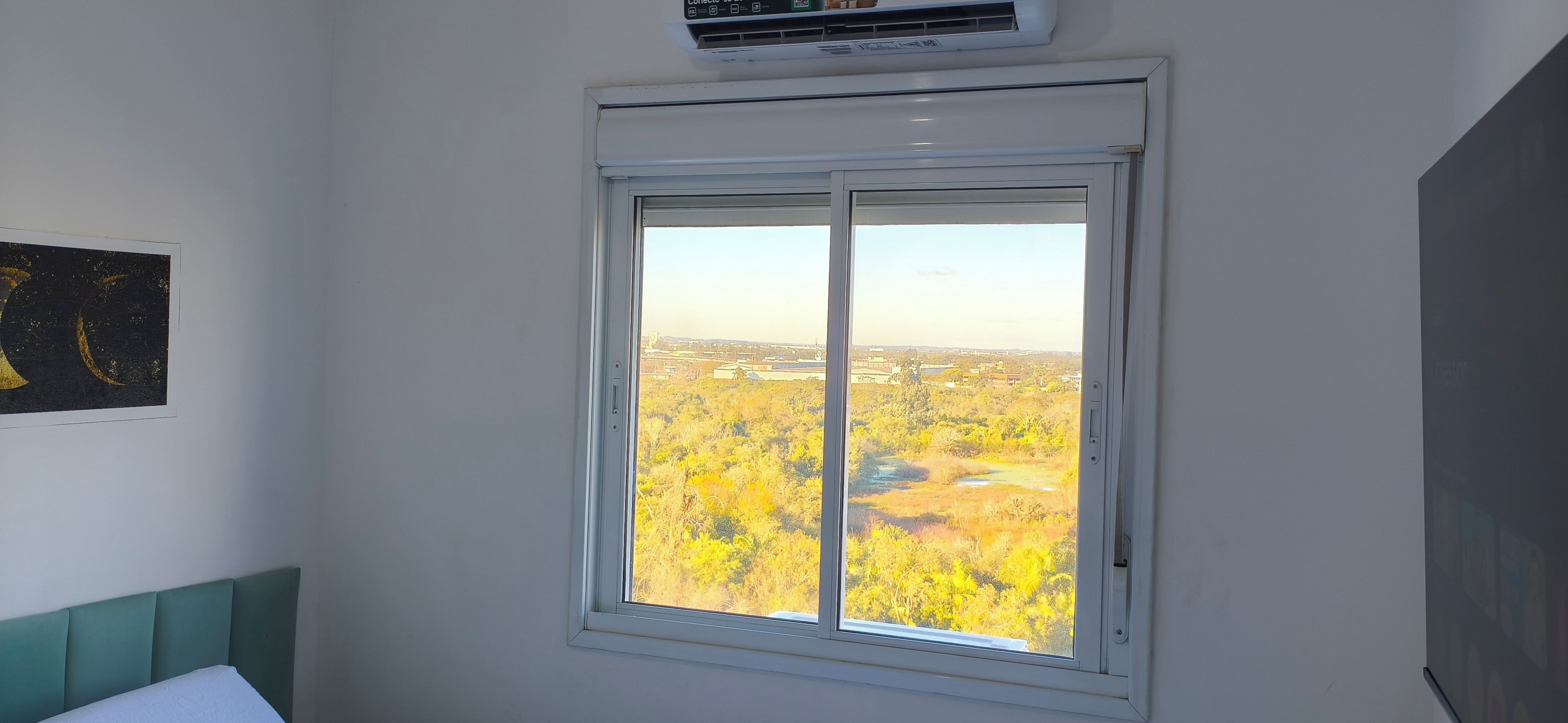 Una habitación con ventana que da a un paisaje, una cama con cabecera verde y un aire acondicionado de pared.