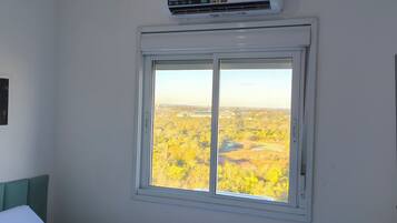 Una habitación con ventana que da a un paisaje, una cama con cabecera verde y un aire acondicionado de pared.