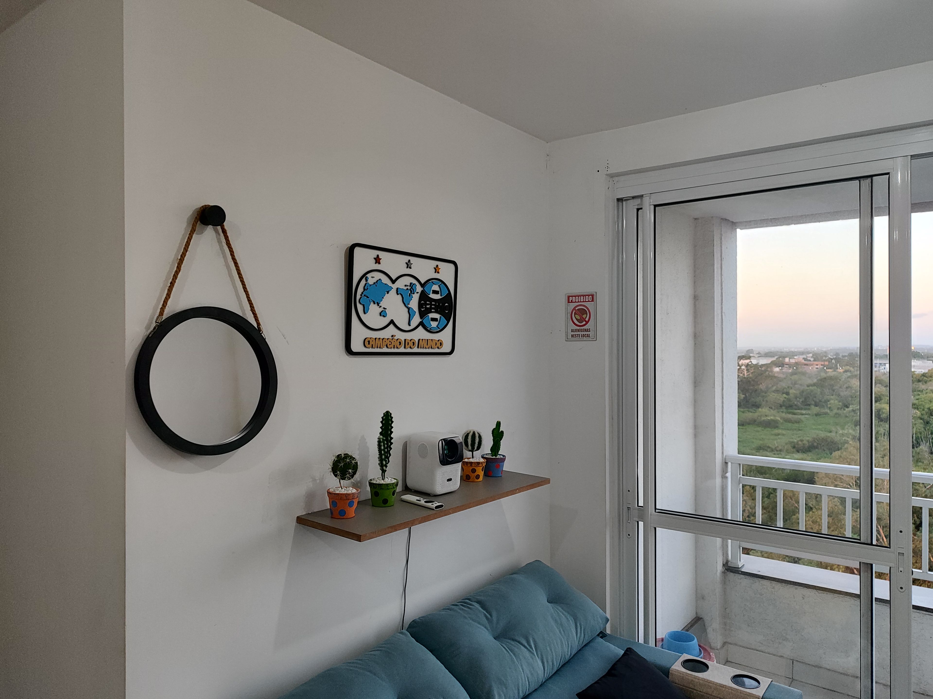 Una habitación con un sofá azul, un estante en la pared con plantas y un electrodoméstico pequeño, un adorno circular y una ventana con vistas a un paisaje urbano.