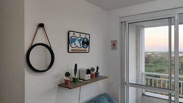 Una habitación con un sofá azul, un estante en la pared con plantas y un electrodoméstico pequeño, un adorno circular y una ventana con vistas a un paisaje urbano.