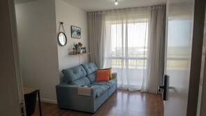 A living room with a blue sofa, a small table, and a window with sheer curtains. - Tricolor Area! Cozy apartment next to Grêmio Arena, near the airport. (Porto Alegre)