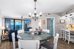 A modern living room with a glass table, white chairs, a blue sofa, and a view of the ocean. - Windward Pointe 3 Bed 2 Bath - Sleeps 10.  Fall Fun! (Orange Beach)