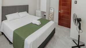 Ein Hotelzimmer mit einem Bett, einer grünen Bettdecke, einem weißen Kopfteil, einem Bild an der Wand, einer Holztür, einem weißen Kleiderschrank, einem weißen Nachttisch, einem Ventilator und einem gefliesten Boden.