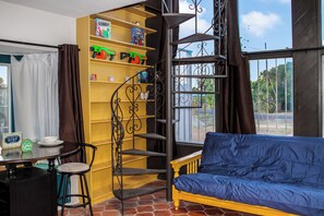 Une piÚce avec un canapé bleu, une étagÚre jaune remplie de jouets, une petite table avec des chaises et un escalier en colimaçon.
