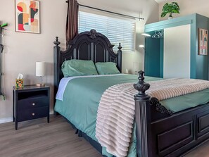 A neatly arranged bedroom with a bed, bedside tables, a nightstand, a potted plant, and a window with blinds.