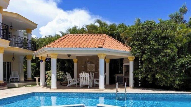 A swimming pool with a tiled deck, a covered seating area with chairs, and a building with a tiled roof.