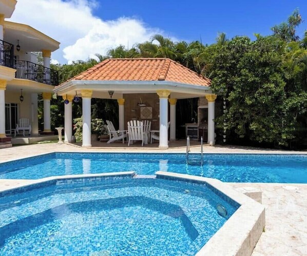 A swimming pool with a tiled deck, a covered seating area with chairs, and a building with a tiled roof. - La Perla by Suarez Properties | Pool, Jacuzzi, BBQ (Juan Dolio)