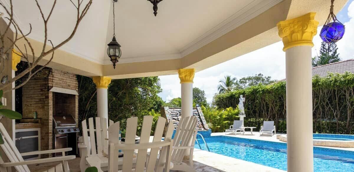 A covered outdoor area with a pool, white chairs, a grill, and a statue.