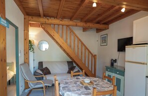 A cozy room with a wooden staircase, a sofa, a dining table with a placemat, and a small kitchen area with a refrigerator. - Riboleys - Maison climatisée à 550m de la mer (Vias)
