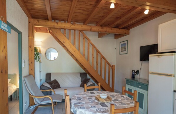 Een gezellige kamer met een houten trap, een bank, een eettafel met placemat en een kleine keuken met een koelkast.