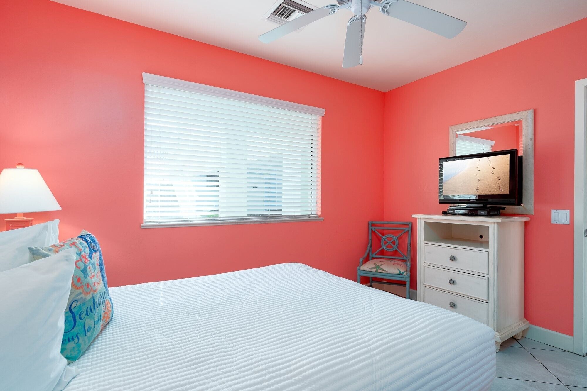 A bedroom with a bed, a television on a stand, a window with blinds, a ceiling fan, and a door.