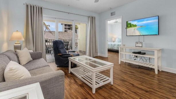 Ein Wohnzimmer mit Sofa, Sessel, Glastisch, Fernseher und einem Balkon mit Blick.
