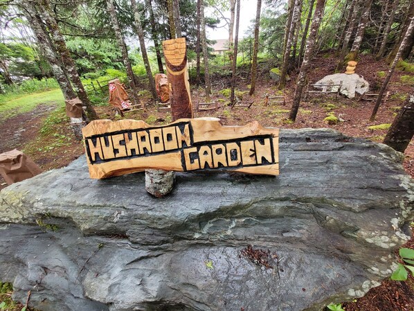 A wooden sign on a rock reading 'Mushroom Garden' in a forested area. - Open-Concept Queen Chalet (Liscomb)