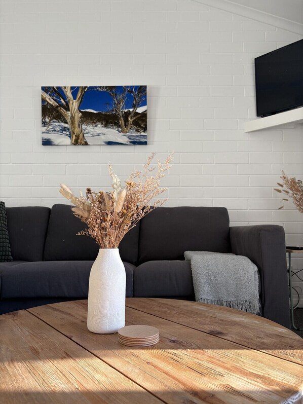 A living room with a dark sofa, a white vase with dried plants on a wooden table, and a painting of trees on the wall.