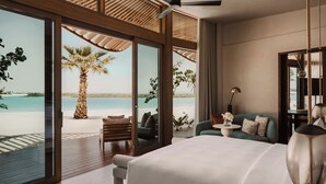 A bedroom with a large bed, a ceiling fan, a view of the beach, and a palm tree.
