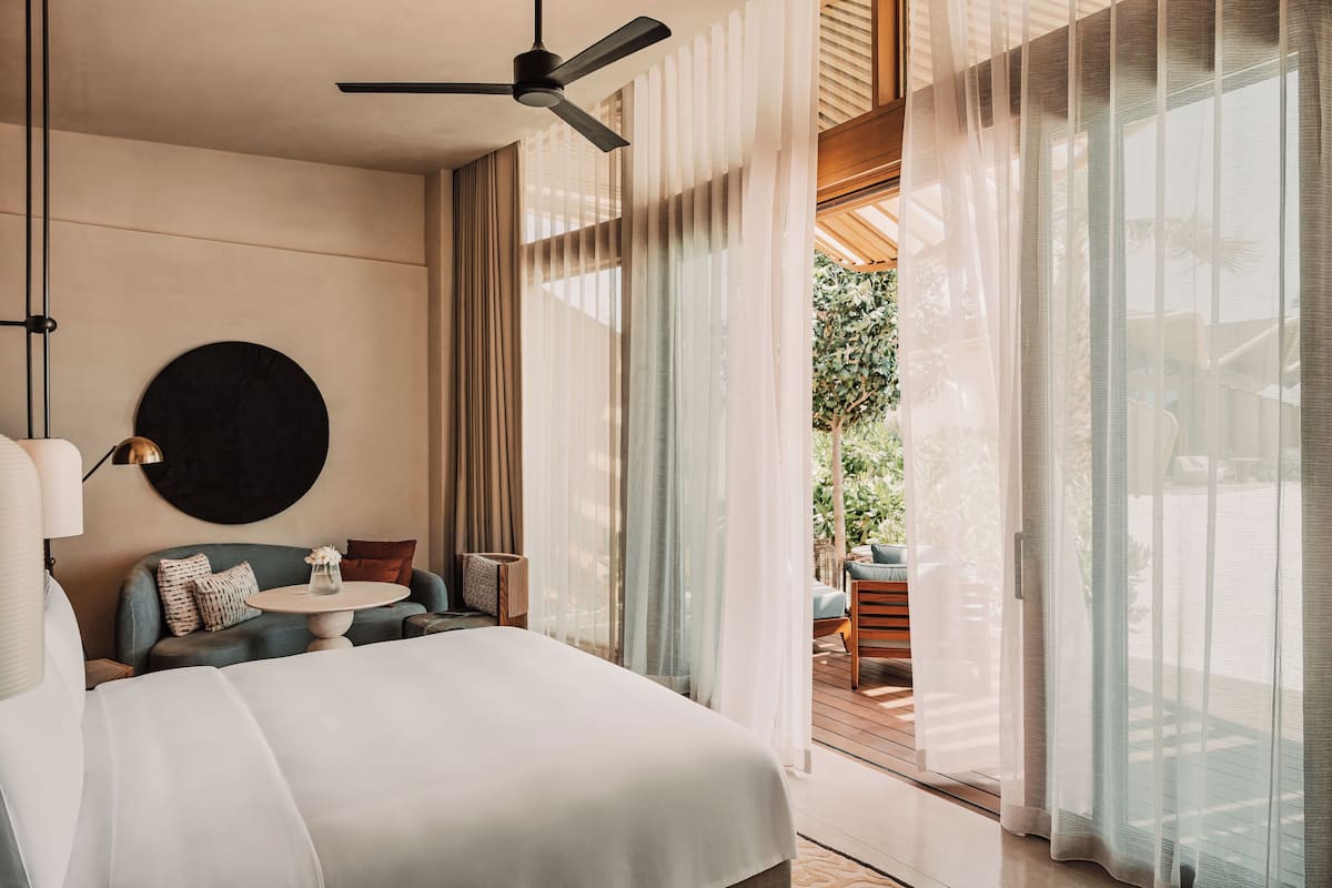 A modern hotel room with a large bed, a round coffee table, a sofa, and a balcony with a view of greenery.