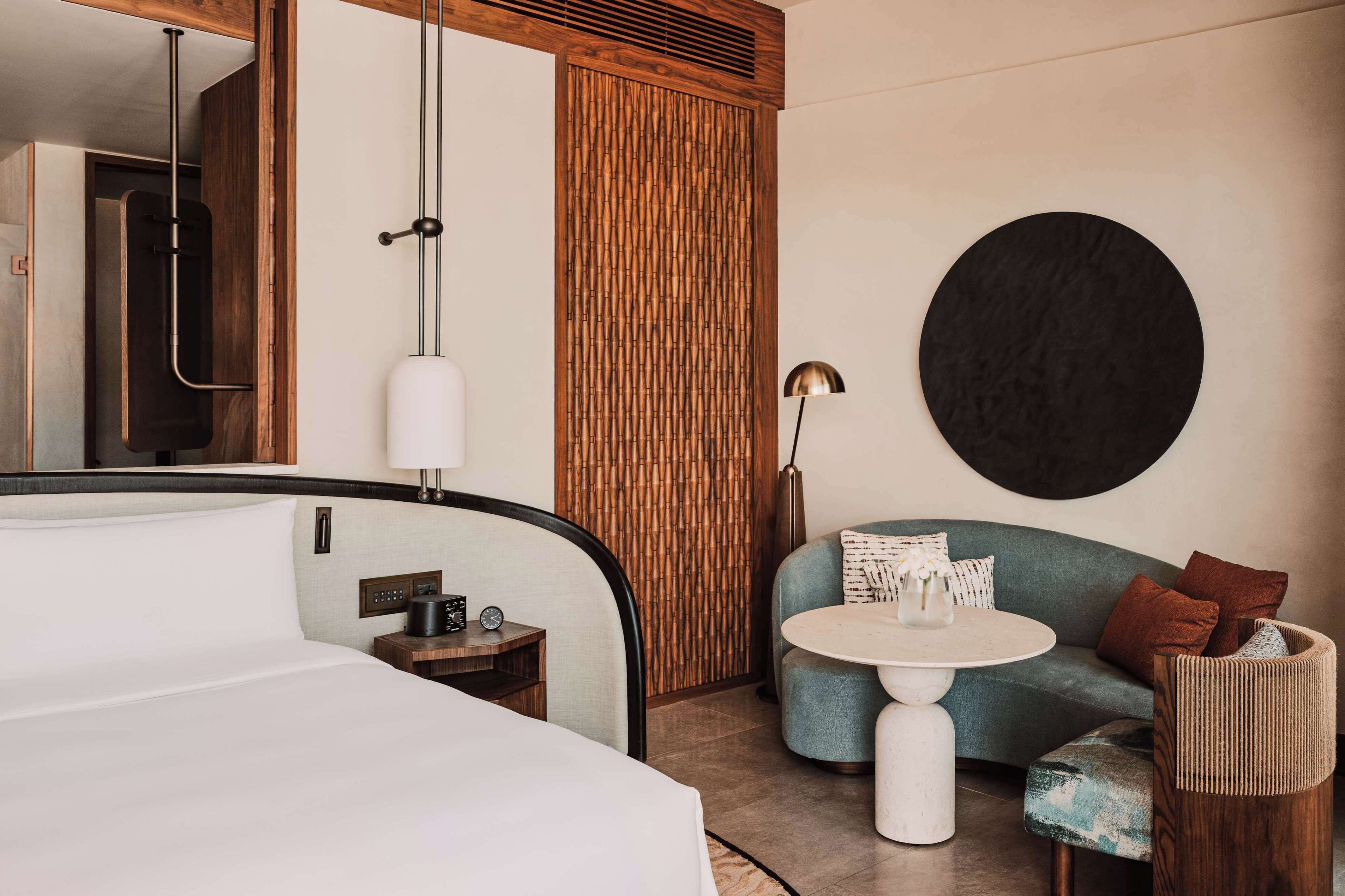 A hotel room with a bed, bedside table, lamp, and a seating area with a table and cushions.