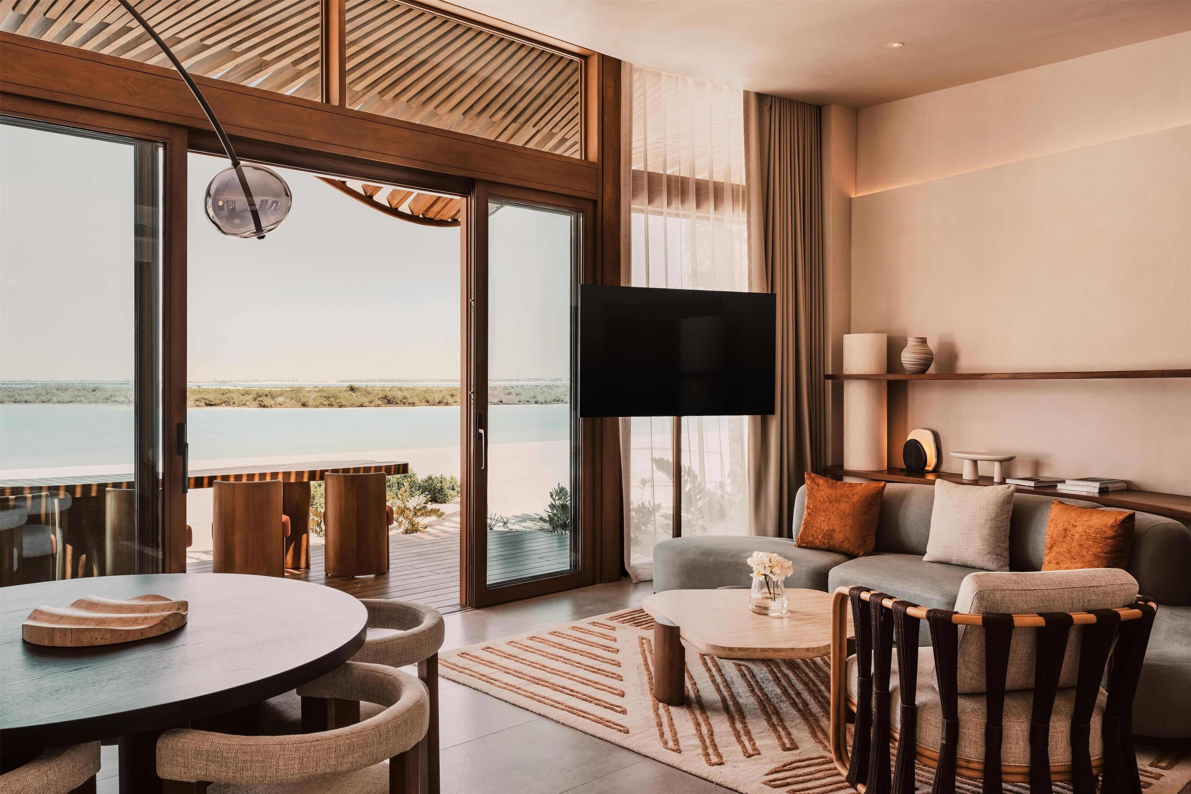 A modern living room with a large window, a flat-screen TV, a round dining table, and a sofa with cushions.