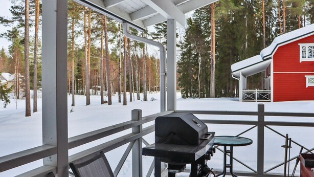 Een overdekt buitengebied met een barbecue, stoelen en uitzicht op een rood gebouw en bomen.