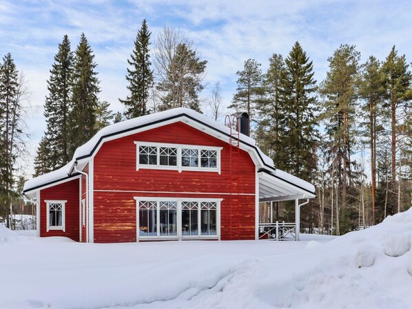A red wooden cabin with a white roof, surrounded by snow and tall pine trees.