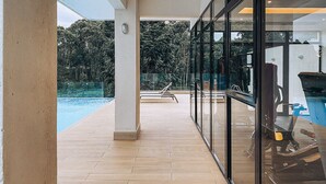 A covered outdoor area with a pool, wooden flooring, and glass walls offering a view of the surrounding greenery. - Luxury 2BR at Two Rivers | Pool, Gym, UN Zone (Nairobi)