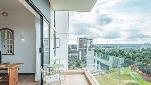 A balcony with a table and chairs, a view of a cityscape, and a glass railing. - Luxury 2BR at Two Rivers | Pool, Gym, UN Zone (Nairobi)