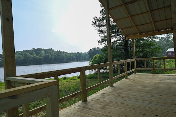 A wooden deck with a railing overlooking a lake, surrounded by trees and a distant house.
