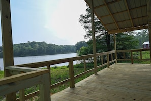 A wooden deck with a railing overlooking a lake, surrounded by trees and a distant house.