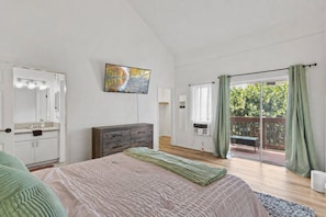 A bedroom with a bed, a dresser, a TV, a balcony with a view of greenery, and a window with curtains.