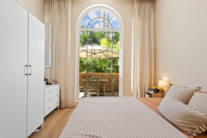 Ein Schlafzimmer mit einem großen Bett, einem Kleiderschrank, einem Schreibtisch mit Lampe und Blick auf eine Terrasse mit Tisch und Stühlen.