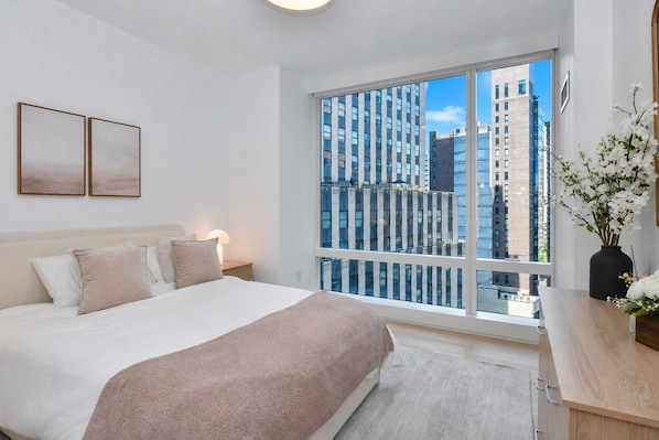 Un dormitorio moderno con una cama grande, una mesita de noche, una lámpara de noche, un ventanal con vistas a la ciudad y un jarrón con flores.