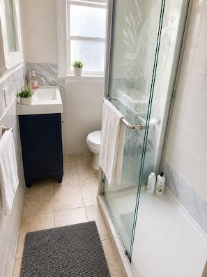 A bathroom with a glass shower door, a white sink, a dark cabinet, a toilet, and a small window with a plant. - Private Rooms Shared Bath UC Berkeley (Berkeley)