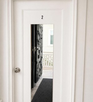 A white door with the number '2' above it, leading to a hallway with a black geometric-patterned door and a view of a white house with a balcony. - Private Rooms Shared Bath UC Berkeley (Berkeley)