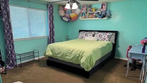 Een slaapkamer met een bed, een bureau, een stoel, een raam met gordijnen en een plafondventilator.