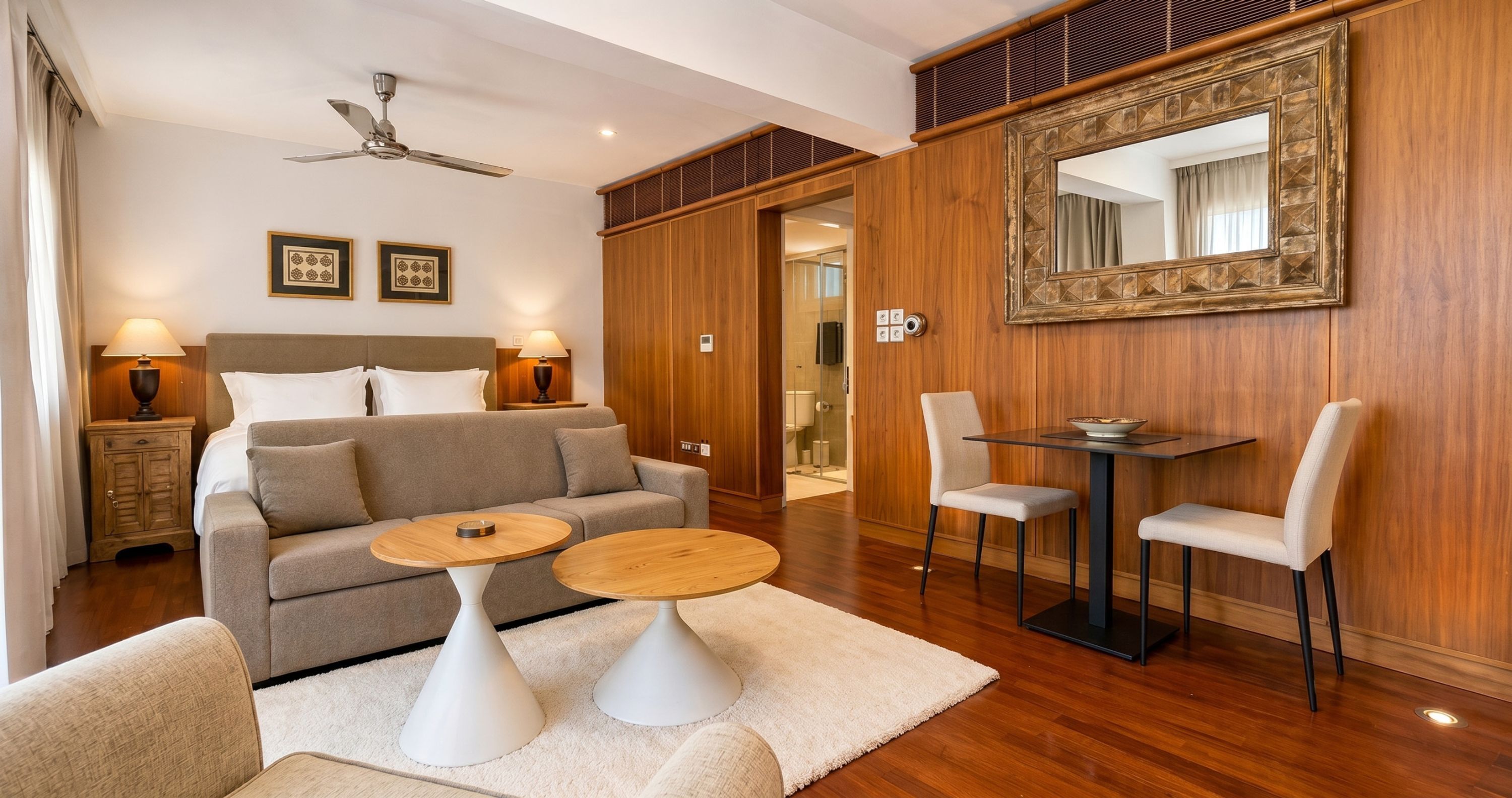 Una camera d'albergo con un letto, due tavolini, un divano, un tavolo da pranzo con sedie, una ventola a soffitto e un grande specchio.