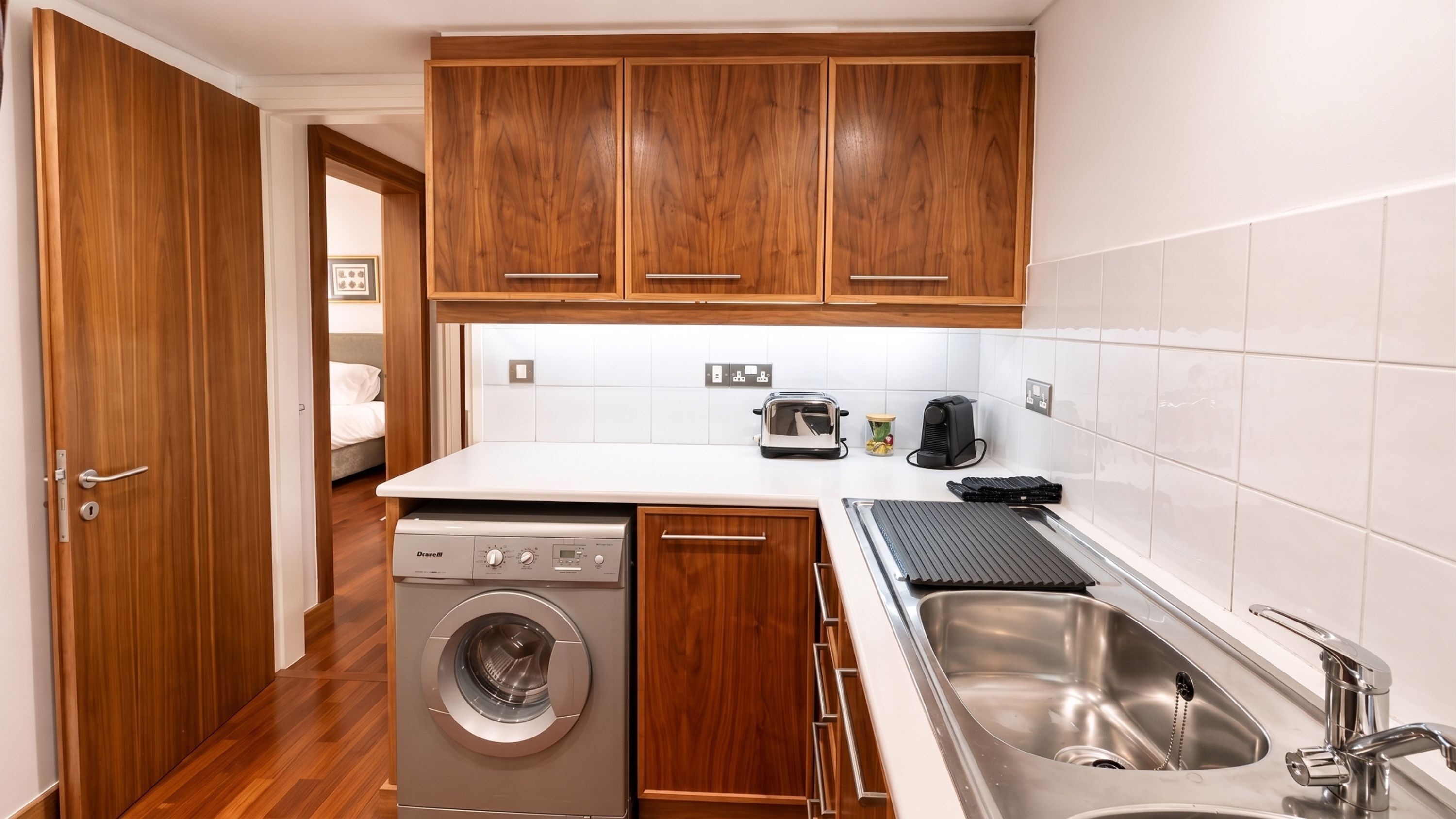 A compact kitchen with wooden cabinets, a built-in washing machine, a toaster, and a sink.