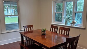 A dining room with a wooden table and chairs, a ceiling light, and windows with views of greenery. - MBC- Cozy home, Direct Access Snowmobile/ATV Trail (Presque Isle)