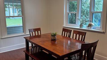A dining room with a wooden table and chairs, a ceiling light, and windows with views of greenery.