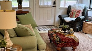 Un salón con un sofá verde, un otoman con estampado, una lámpara, una silla con cojín blanco y una puerta con cortina blanca.
