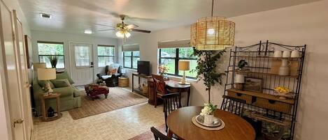 A dining area with a round table, chairs, a hanging light fixture, and a wall-mounted shelf with dishes.