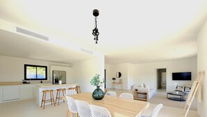 A modern kitchen with white cabinets, a dining area with a wooden table and white chairs, a living room with a sofa and a TV, and a hallway leading to another room. - Villa Di Luna Di San Ciprianu (Zonza)