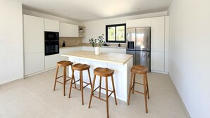 A modern kitchen with white cabinetry, a central island with stools, a stainless steel refrigerator, and a window providing natural light. - Villa Di Luna Di San Ciprianu (Zonza)