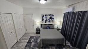Une chambre d’hôtel comprenant un lit, deux tables de chevet, une œuvre d’art murale, un luminaire au plafond et une fenêtre avec des rideaux.