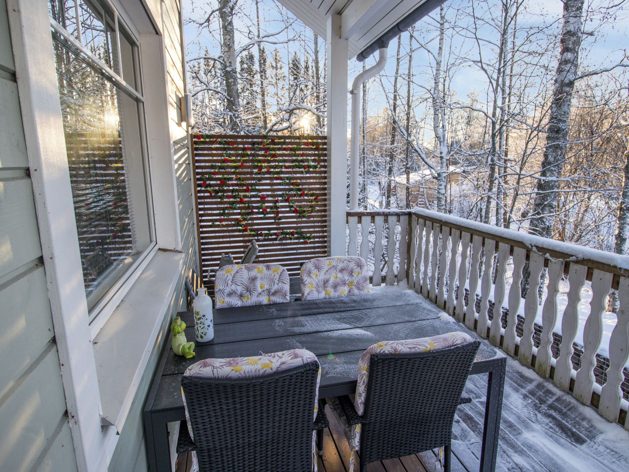 Katettu kuisti, jossa on pöytä ja tuoleja, koristeellinen aita ja näkymä lumipeitteisiin puihin.