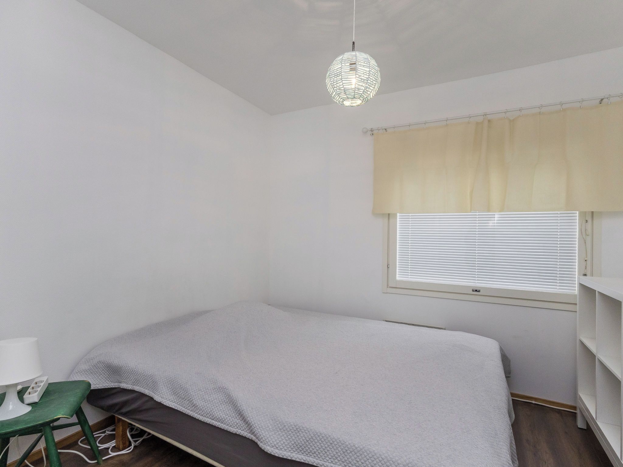 A small, single-bed room with a desk and chair, a window with blinds, and a hanging light fixture.